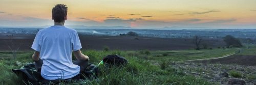 man sits on green grass facing backward meditating in sunset dark sky