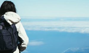 woman facing backward wearing white jacket black back bag stands looking at blue cloudy sky