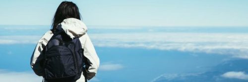 woman facing backward wearing white jacket black back bag stands looking at blue cloudy sky