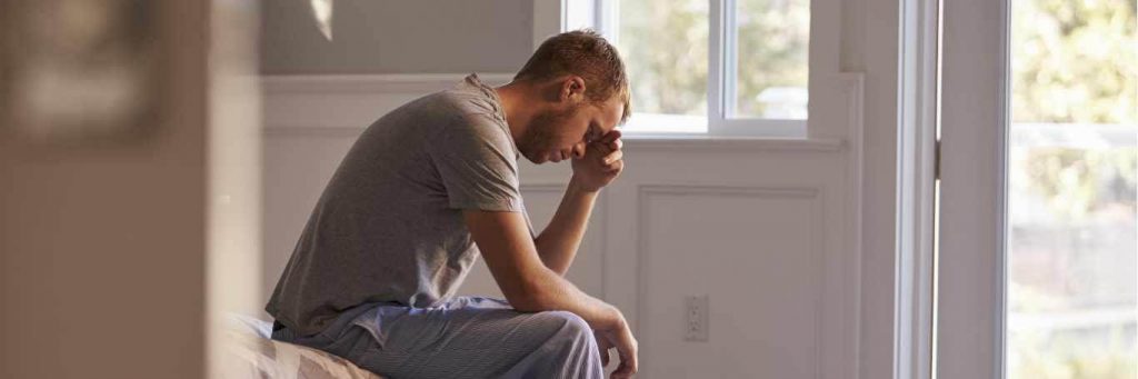 man sits on bed hand in face sadly thinking during challenging period