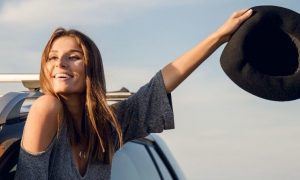 woman stands raising hand with black hat from car window happily smiling