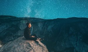 man sitting on on rock watching blue star sky