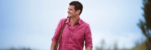 man wearing side bag stands on field smiling in blue clear sky
