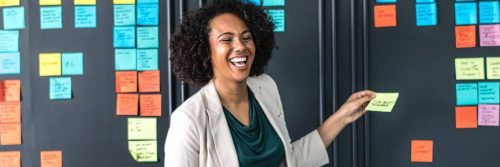 curly black hair woman stands in classroom happily laughing putting yellow stick note on black board