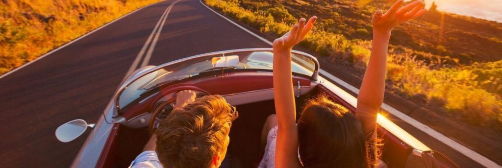 couple sits in car driving on empty road while woman excitedly raising hands in beautiful sunny sky