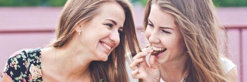 two women sits on bench talking laughing while one lady eating biscuit