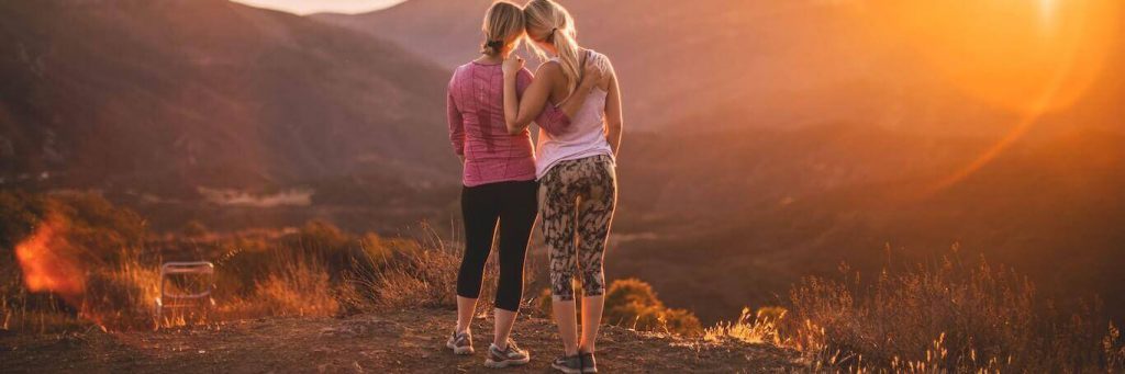 two women stand on rock side by side facing backward enjoying beautiful sunny weather