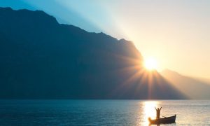 person stands on boat raising hands bloating on blue ocean approaching mountain in blue sunny sky