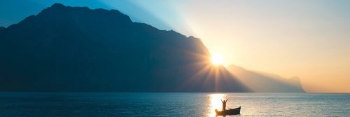 person stands on boat raising hands bloating on blue ocean approaching mountain in blue sunny sky