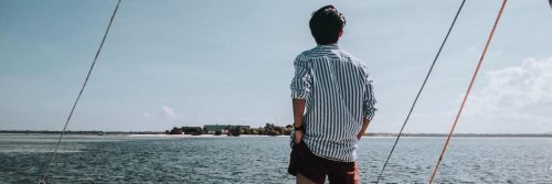 man facing backward hands in pocket stands on boat looking at blue ocean island in cloudy sky
