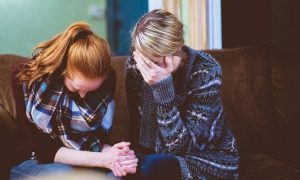 young woman sits on couch face down crying beside mom sitting beside hand in face crying