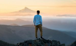 man facing backward stands on rock hands in pocket looking at mountains