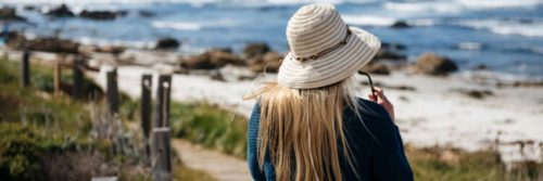 woman facing backward walking along coastal walk beside blue ocean rocks