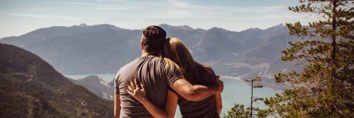 couple facing backward sits on mountain top hugging while woman lying on boyfriend shoulder watching cloudy sky