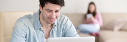 man smiles studying laptop while woman sitting using mobile phone on white couch