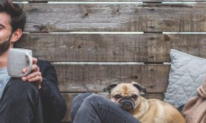 man sits beside wall holding grey mug smiling while woman sitting smiling at puppy