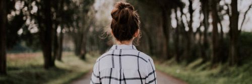 woman facing backward stands on road between forest