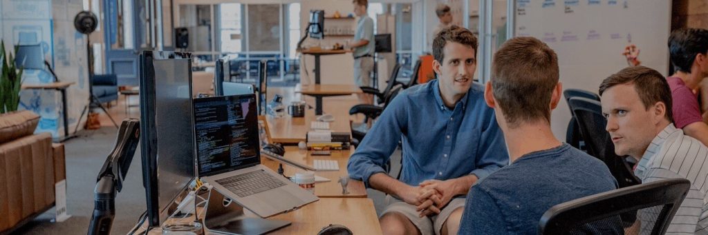 three men sits in busy laboratory besides laptops computers chatting