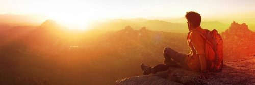 man sits on mountain top wearing back bag watching sunset