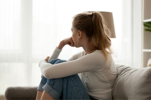 Side view young woman looking away at window sitting on couch at home filled with loneliness 