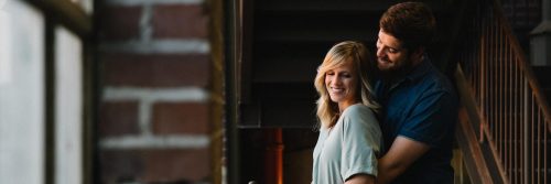 man hugs girlfriend from behind while woman happily smiling standing beside stair