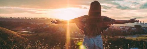 woman facing backward stands on field raising hands gratitude life in peaceful environment in sunny sky