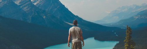 man stands beside lake looking at mountain in foggy weather