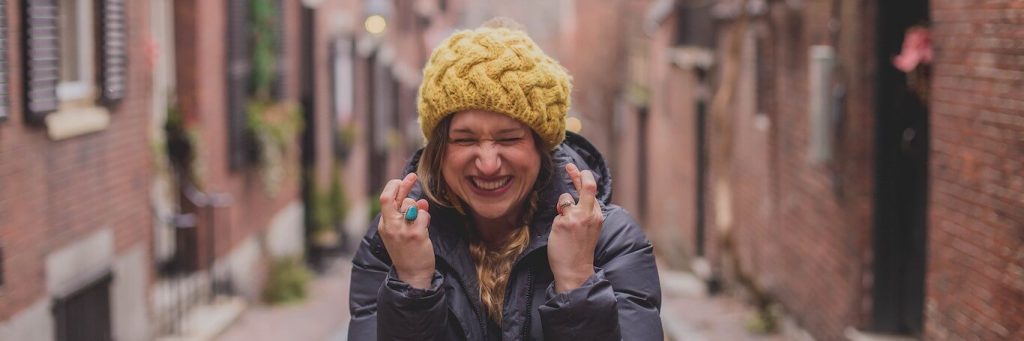 excited woman with yellow beanie and dark blue winter coat stands alone between two arrays of buildings