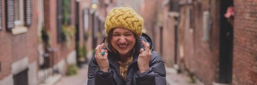 excited woman with yellow beanie and dark blue winter coat stands alone between two arrays of buildings