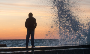 man stands on seaport looking at strong ocean wave in sunset sky